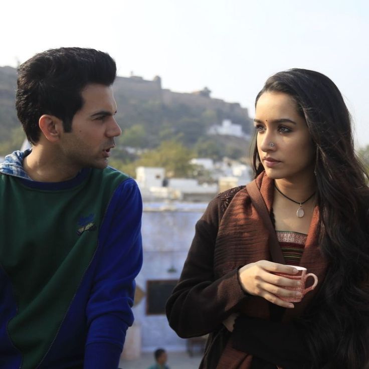 This may contain: a man and woman standing next to each other looking at something in the distance with buildings in the background